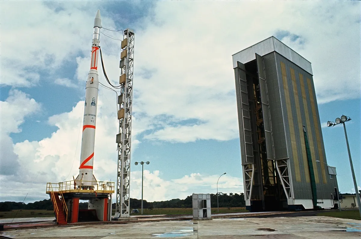 Colour photo of a white Diamant B rocket with red stripes on its launch table beside an umbilical mast, with the service tower rolled back, at the Guiana Space Centre in 1970